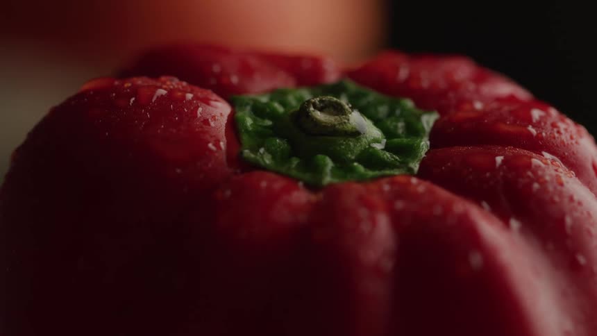 A dramatic extreme closeup shot focuses on the green stem of a vibrant red bell pepper its glossy skin covered in fine droplets of water against a completely black background