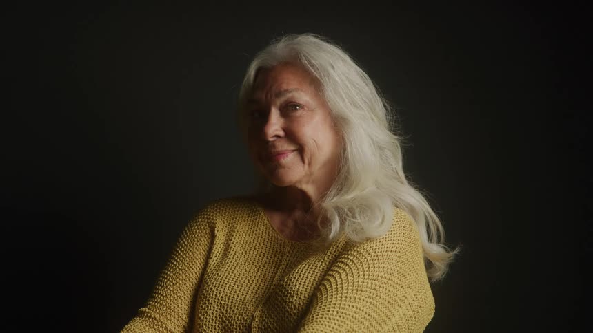 An elderly woman sits in a dark room wearing a yellow sweatshirt smiling