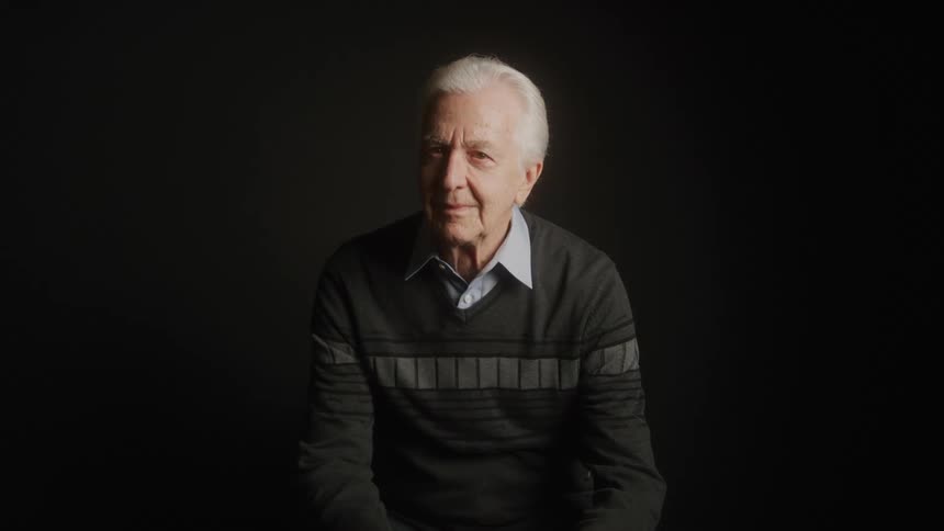 The video shows a closeup of an elderly man with white hair wearing a striped grey and black sweater he is looking directly at the camera with a neutral expression the background is a solid dark color
