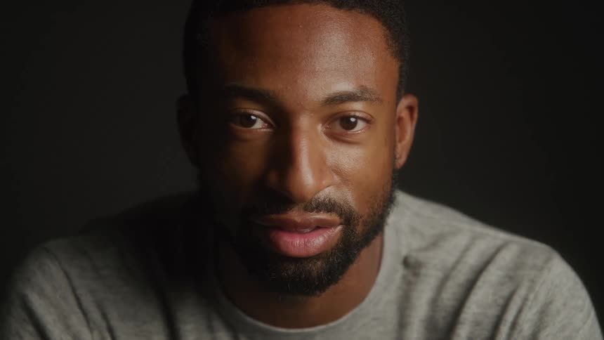 A black man with a trimmed beard and calm expression gazes directly into the camera under soft focused lighting