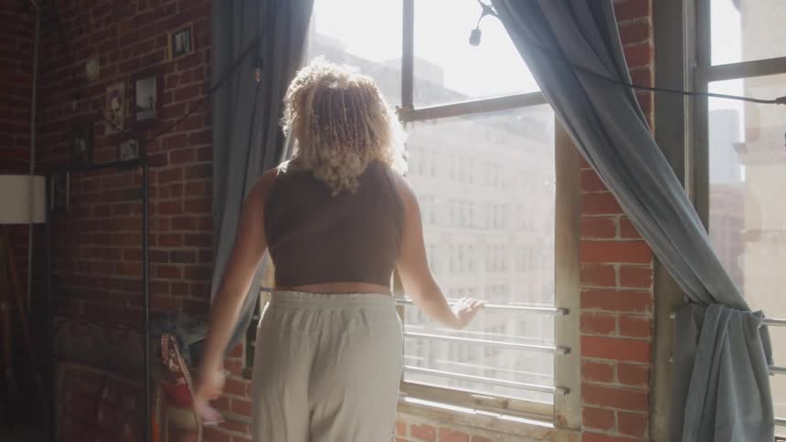 A young woman resting her hands on a glass window indoors looking sad then slowly walking toward a chair