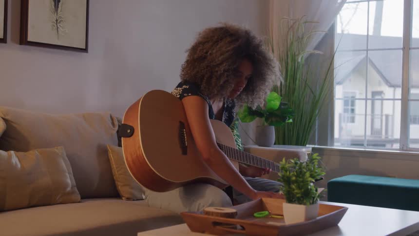 A woman sits on her couch while practicing a song on her guitar