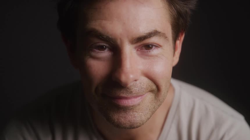 A man with dark hair and stubble smiles subtly while looking directly at the camera with a dark background