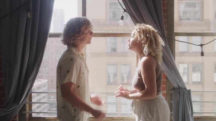 A couple argues in a sunlit apartment standing face to face by the window