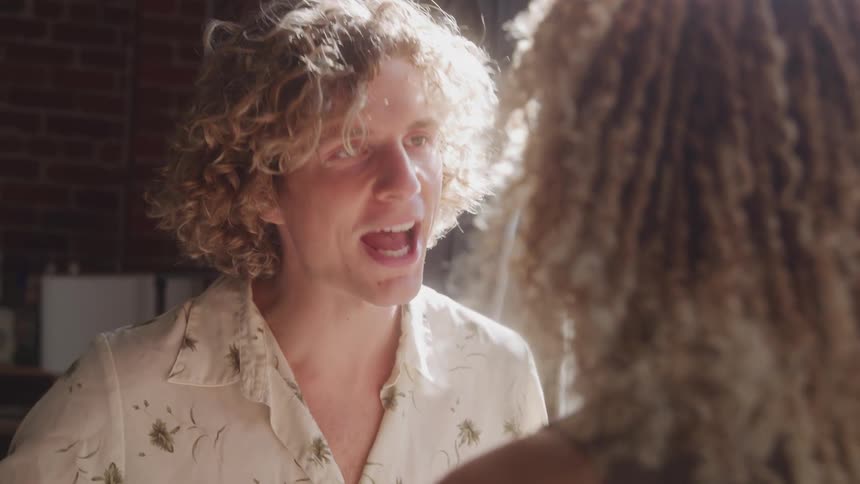 A man and a woman with curly hair stand next to a window indoors engaged in a heated argument