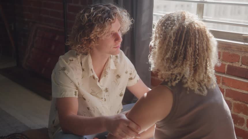 Two individuals with curly hair are facing each other seemingly engaged in a deep conversation one person wears a lightcolored patterned shirt while the other seen from behind has very light blonde or gray curly hair