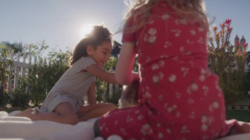 Two young girls sit on a light colored blanket outdoors under bright sunlight with one girl in a grey shirt and the other in a red patterned dress as they play with a small stuffed dog between them