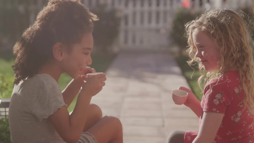 Two young girls are sitting facing each other outdoors appearing to be having a tea party the girl on the left has dark curly hair and is wearing a grey tshirt while the girl on the right has long blonde hair and is wearing a red floral dress