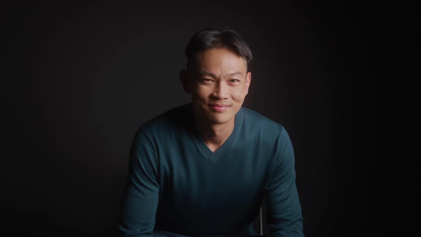 A man in a teal sweater smiles warmly while seated against a dark softly lit backdrop