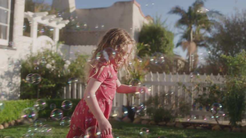 A young girl with curly blonde hair wearing a red patterned dress playing with floating bubbles in a sunny backyard with a white picket fence and house in the background
