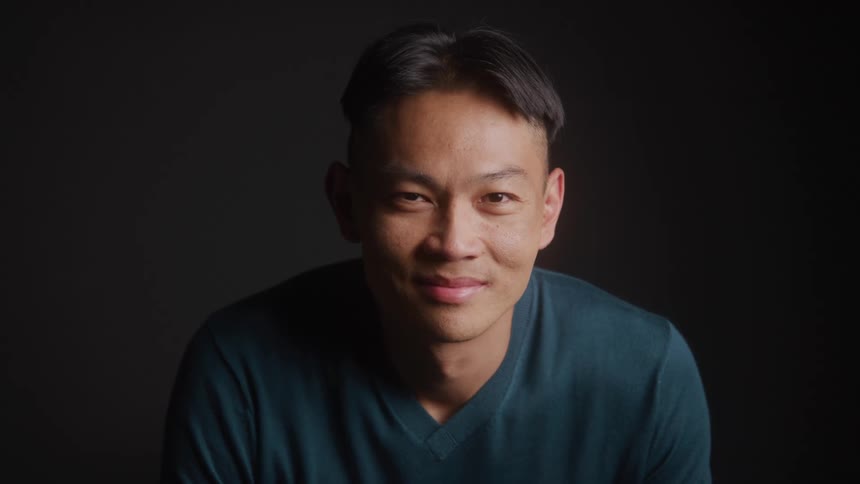 A closeup shot features a man with short dark hair and a faint smile looking directly at the camera he is wearing a dark teal or blue vneck shirt against a dark background with soft lighting highlighting his face