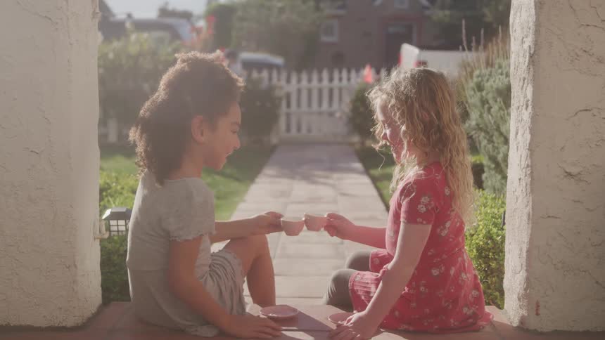 Two children sit on the front porch of a house and cheers with tea cups