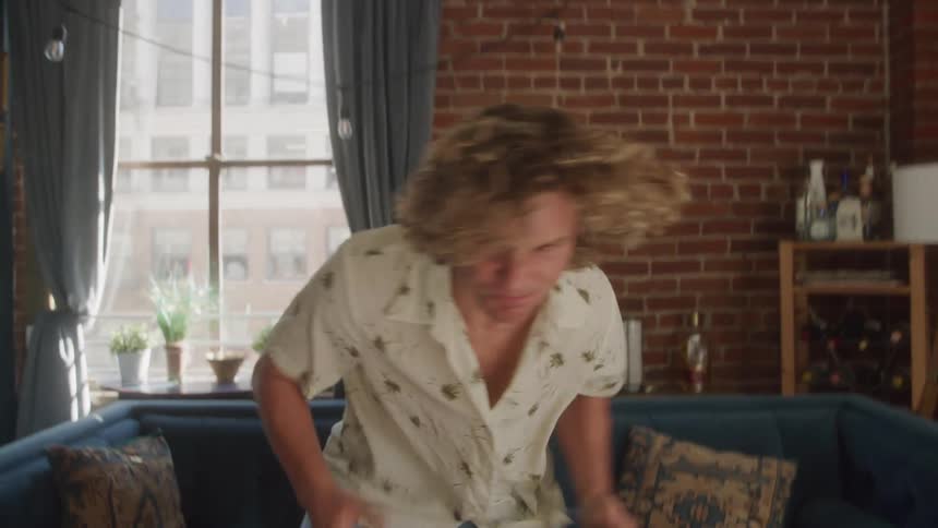 A person with curly blonde hair wearing a patterned shirt walks towards a blue couch in a room with a brick wall and a large window then sits down and reacts with a grimace