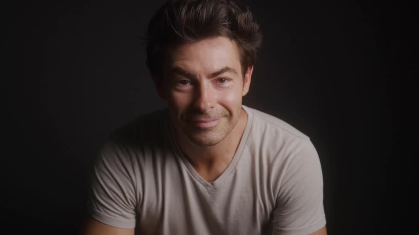 A smiling man with tousled hair and a light tshirt sits against a dark background