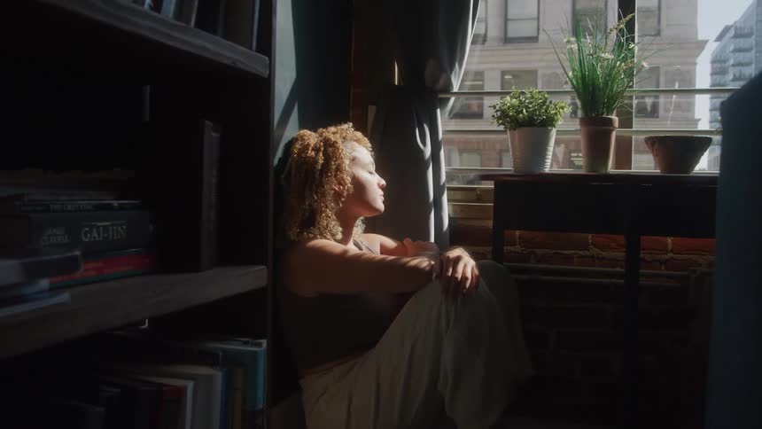 A young woman sitting on the floor indoors lost in thought with a potted plant on a table in front of her at midday