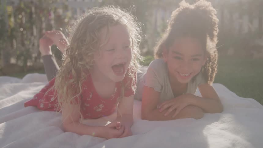 The video features two young girls one with curly blonde hair and the other with curly dark hair lying on a white blanket outdoors in what appears to be a sunny setting the blondehaired girl is crying with her mouth wide open while the darkhaired gi