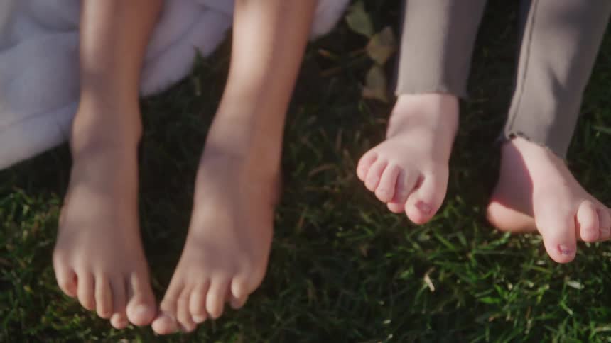 Two kids sit on a blanket laid on a lawn at midday moving their bare toes up and down