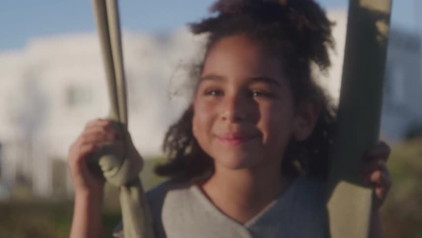 The video shows a young girl with curly hair on a swing smiling directly at the camera the background appears bright and sunny with hints of a building and clear sky