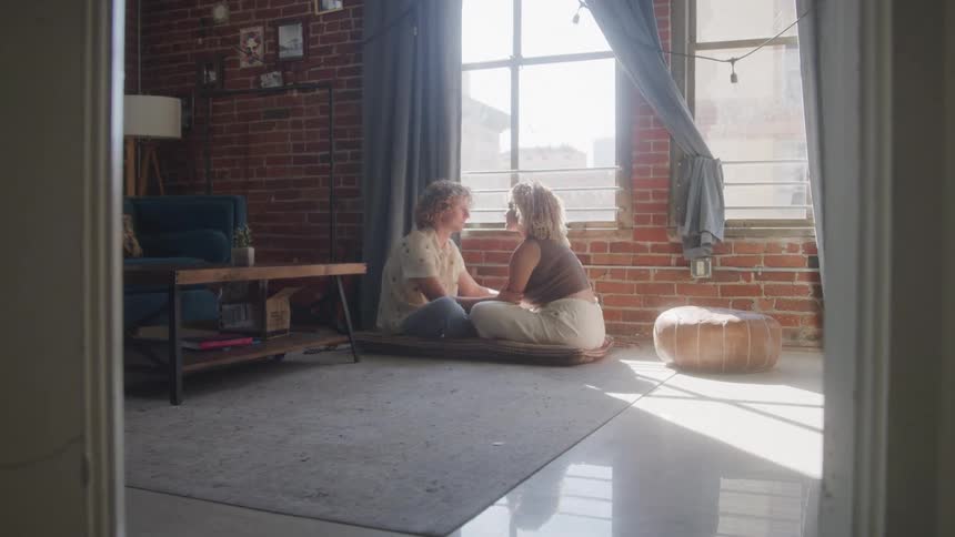 A couple sits on the floor gazing into each others eyes in a sunlit living room