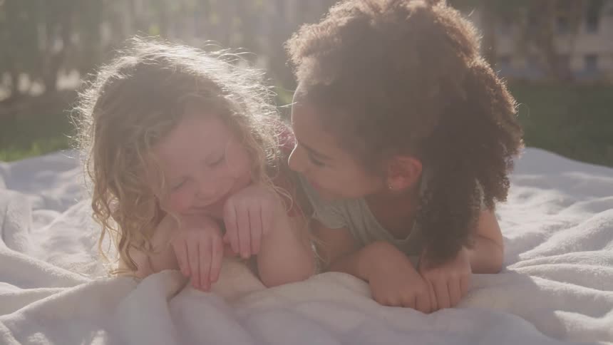 Two young girls with different hair colors are lying on a white blanket on a grassy area looking up at the camera they are smiling with bright sunlight creating a halo effect around their heads
