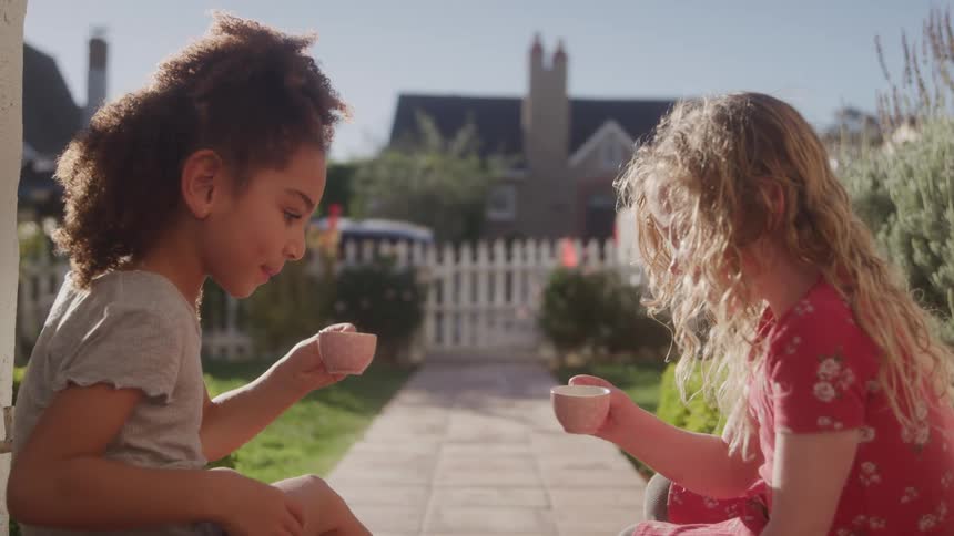 Two young girls are sitting outdoors facing each other one girl with curly dark hair is on the left and another with long blonde hair is on the right both holding small teacups as if having a tea party in the background a house and a white picket fe