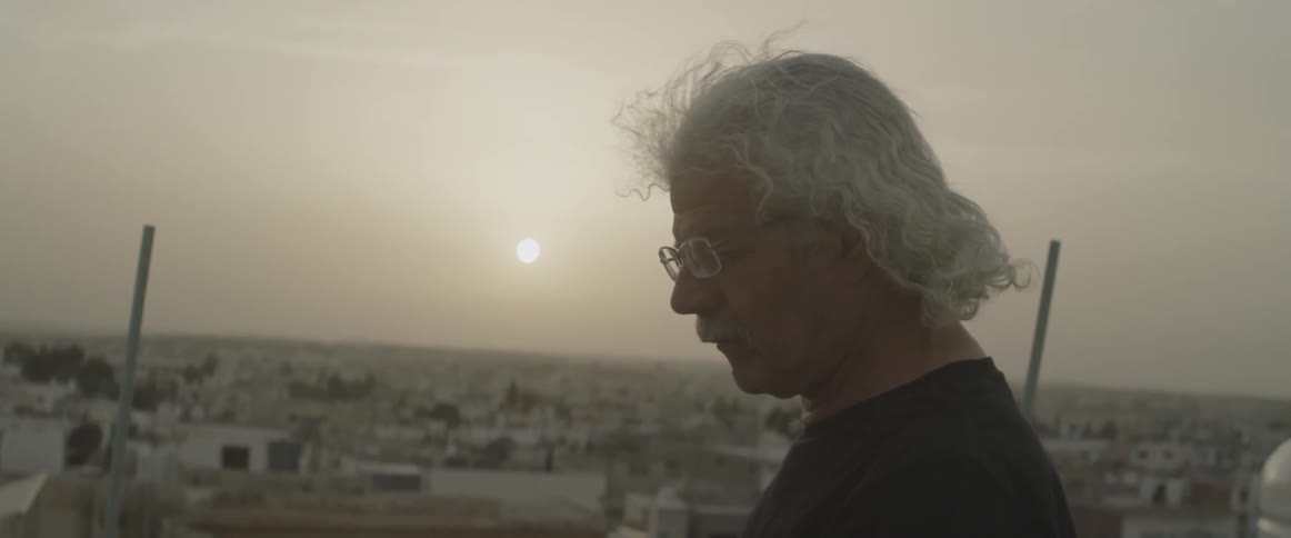 A man with curly gray hair looks down near sunset