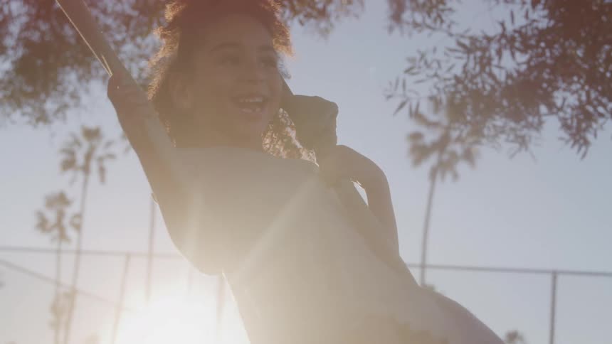 A happy young girl with curly hair playing on a swing rope on a sunny day and a backdrop of palm trees and a chainlink fence