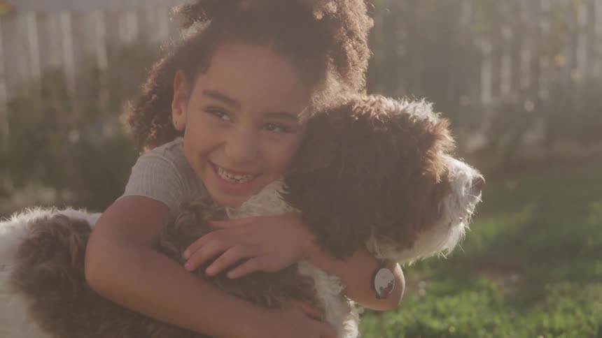 A young girl playfully hugs her dog on the lawn outdoors during daytime