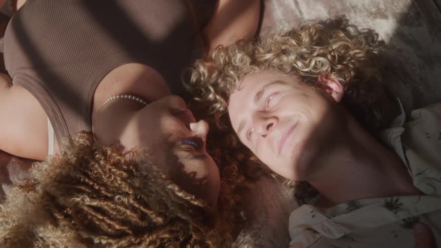 The video shows two individuals one with dark dreadlocks and the other with curly blonde hair lying down next to each other sunlight falls across their faces creating a warm and intimate atmosphere