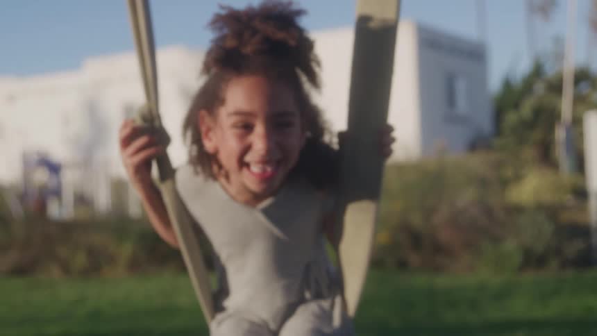 A joyful young girl with curly hair wearing a gray tshirt playing on a swing rope on a bright sunny day