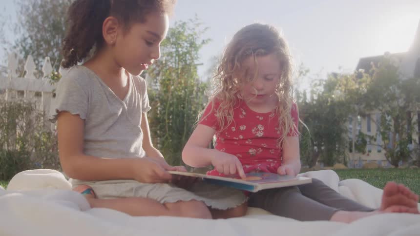 Two kids sit on a blanket on the lawn playing with a storybook at midday