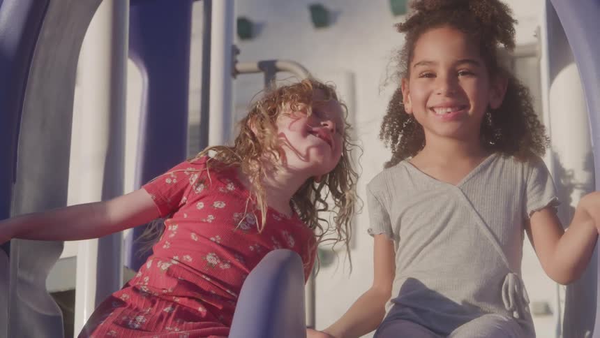 Two young girls one with blonde curly hair and a red floral dress and the other with dark curly hair and a grey top are sitting on a blue and white playground slide they are looking at each other and smiling appearing to be enjoying themselves on a s