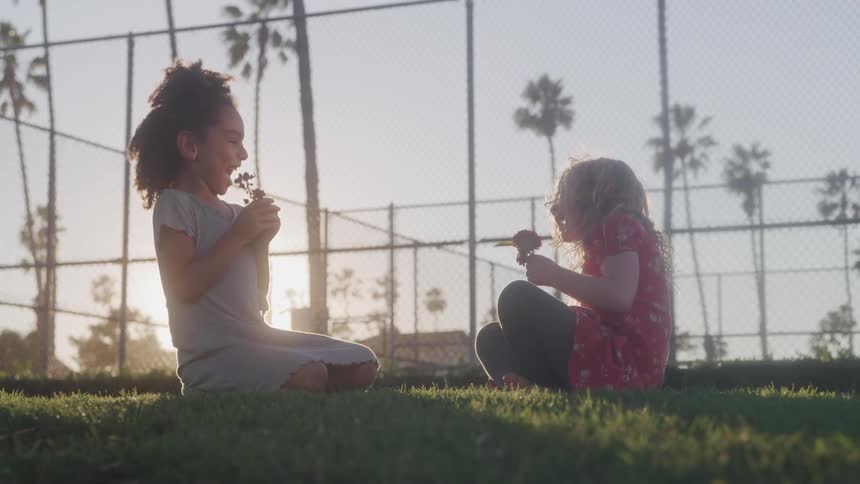 Two girls sit on grass at sunset laughing and holding flowers