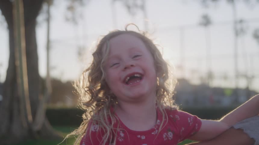Two young children with the sun brightly backlighting them are seen smiling and laughing outdoors one child has long lightcolored curly hair and is wearing a red patterned top
