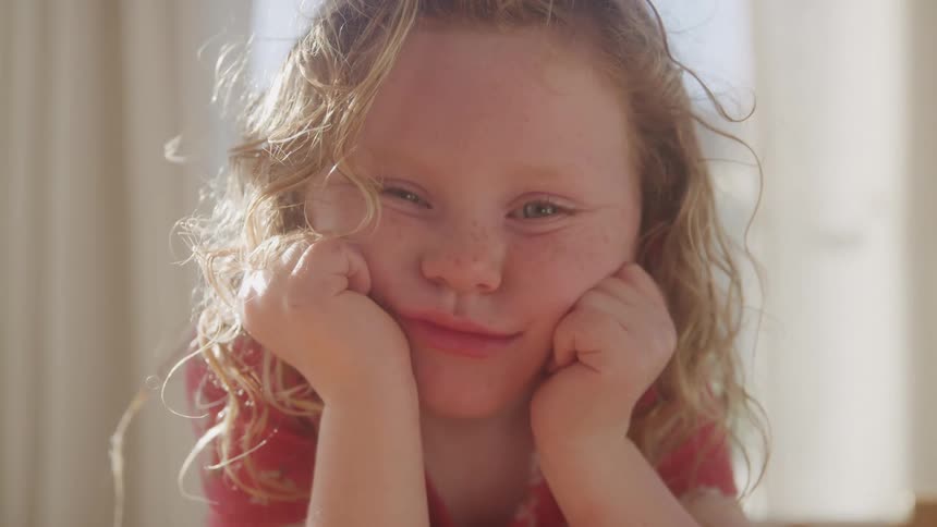 A young girl with curly blonde hair rests her chin in her hands smiling softly in warm sunlight