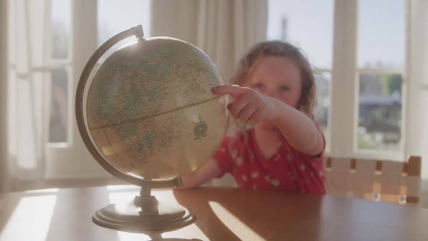 The video features a young girl with curly blonde hair wearing a red shirt seated at a wooden table she is actively engaged with a globe pointing at different locations and seemingly exploring it