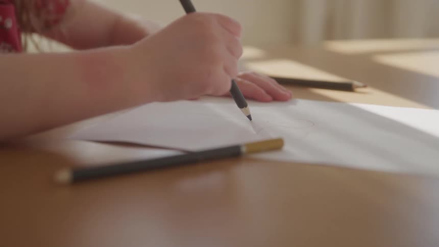 A child draws with a pencil on paper at a sunlit desk