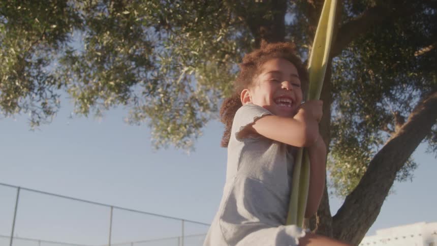 A young girl with curly hair is joyfully swinging from a yellow rope attached to a large tree she is smiling broadly and appears to be having fun in a parklike setting with other children in the background