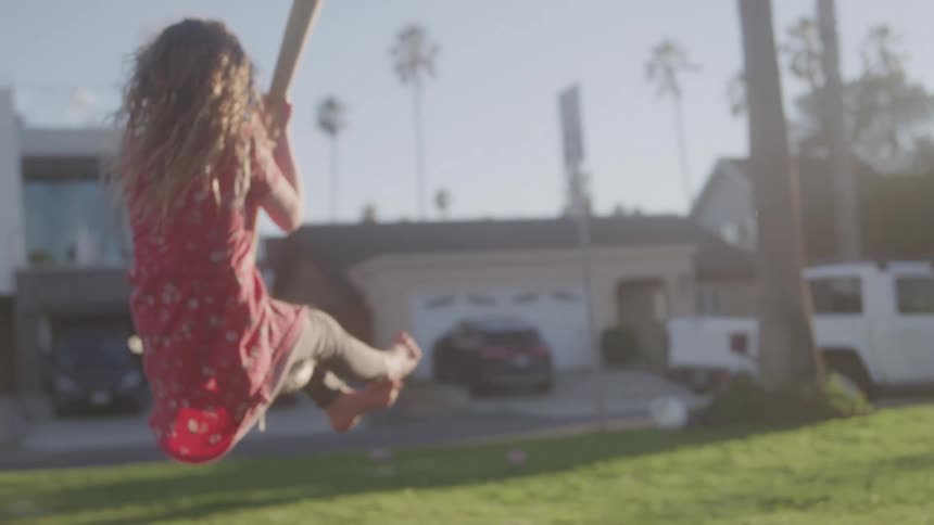 A young girl with long curly blonde hair is joyfully swinging on a swing set in a sunny outdoor setting her red and white patterned dress is visible as she swings high with houses and palm trees in the background