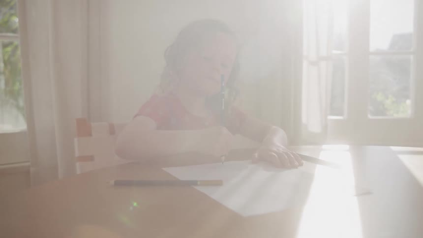A young girl sits at a dining table writing on paper with a pencil indoors on a sunny day