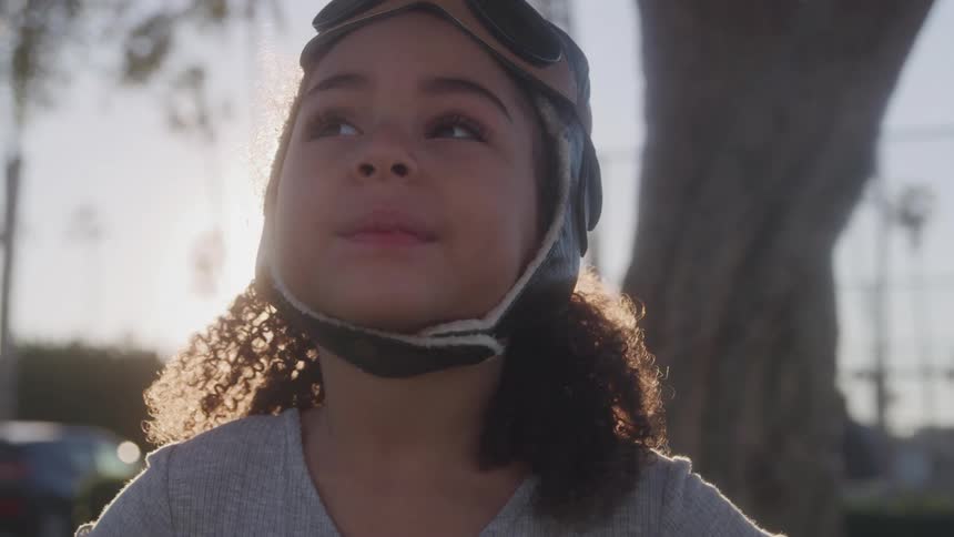 A young girl wearing an aviator hat with goggles is seen in a closeup shot looking upwards with a thoughtful or hopeful expression the sun is shining brightly behind her creating a halo effect around her curly hair