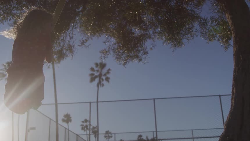 A person with long flowing hair is silhouetted against a bright sky swinging from a rope or swing attached to a large tree in the background palm trees and a chainlink fence are visible