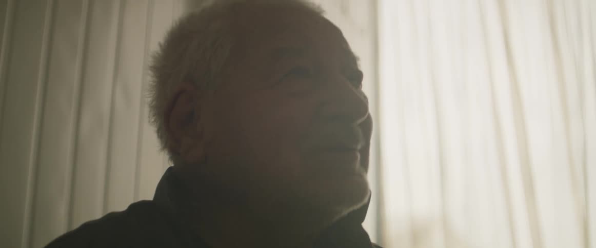 An old man sits by a window smiling and talking