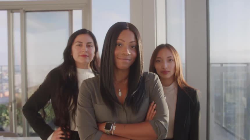 Three confident businesswomen stand in a sunlit highrise office exuding professionalism and unity as they face the camera with poised expressions