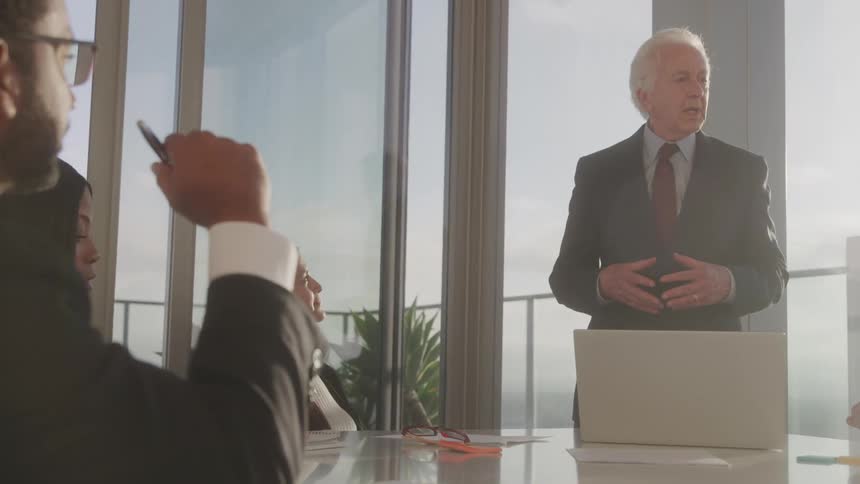 An elderly man gives a presentation in a conference room with his coworkers at midday