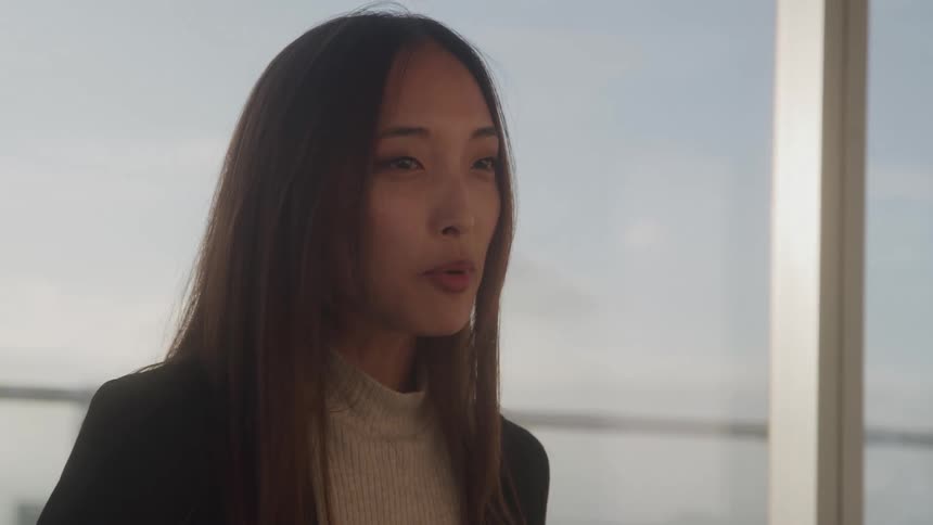 A young asian woman with long straight dark hair and a black blazer speaks animatedly her expressions changing from open mouthed talking to a slight smile as seen against a bright out of focus window background