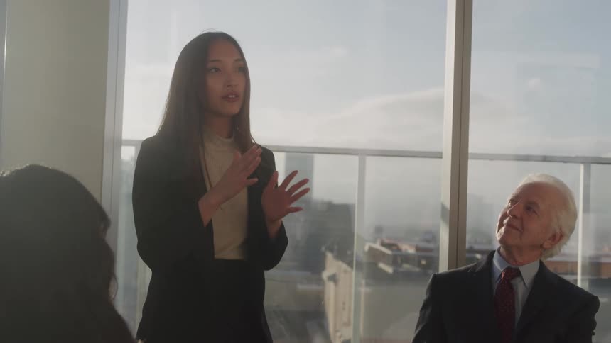 A woman in a black blazer and beige top is speaking gesturing with her hands in what appears to be a business meeting an older man in a suit looks up attentively from the right side of the frame while another persons back is visible on the left
