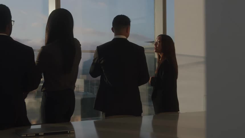 A group of business professionals are gathered in a modern office standing by a large window with a city view they are silhouetted against the bright light coming through the window