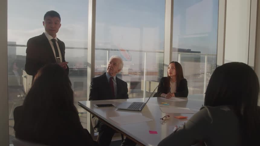A young male businessman stands while speaking to his colleagues in a meeting room during daytime
