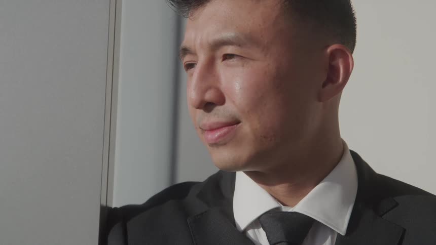 An asian man in a black suit and tie gazes thoughtfully out a window bathed in natural light